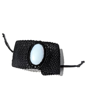 May include: Black macrame bracelet featuring a light blue oval stone centerpiece. The bracelet has a woven texture and is secured with black cords. A stylish accessory for everyday wear.