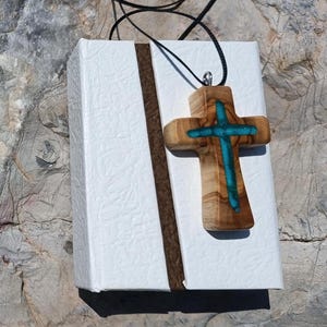 Puede incluir: Colgante de cruz de madera con incrustaciones turquesas, suspendido de un cordón negro. La cruz se muestra sobre una caja de regalo blanca con una franja marrón. El fondo es una superficie de piedra clara y texturizada.