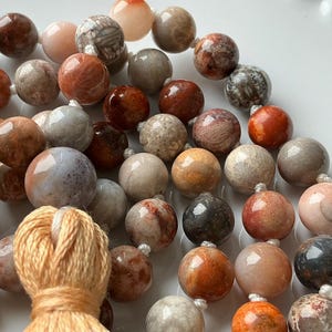 May include: Close-up of a beaded necklace featuring a variety of earth-toned, round beads. The beads range in color from light pink to dark brown, with some displaying unique patterns. A tassel of orange thread is visible at the bottom.