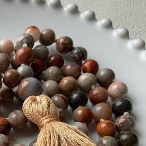 May include: A close-up of a beaded necklace with a tassel. The beads are various shades of brown, gray, and pink. The tassel is a light tan color. The necklace is resting on a white, scalloped surface.