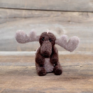 May include: A brown felt moose ornament with large, grey felt antlers. The moose is sitting on a wooden surface.