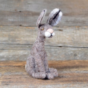 May include: A hand-felted gray rabbit with white fur on its ears, sitting on a wooden surface. The rabbit has brown eyes and a pink nose.