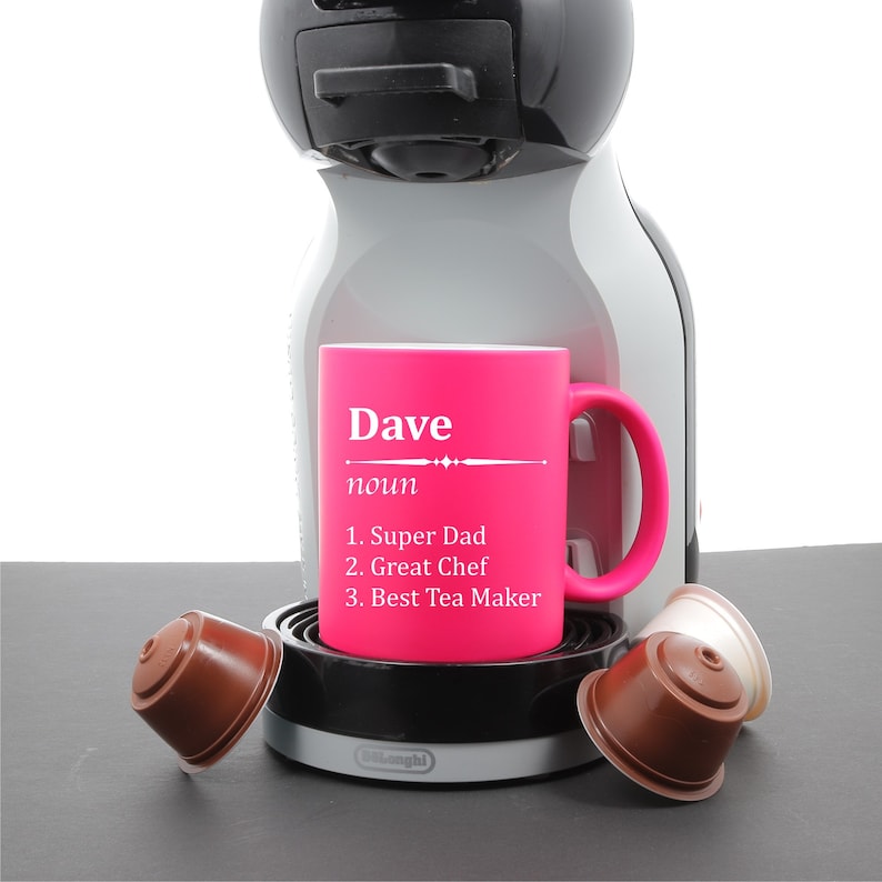 May include: Pink coffee mug with the word "Dave" and the definition "noun 1. Super Dad 2. Great Chef 3. Best Tea Maker" printed on it. The mug is sitting on a coffee machine.