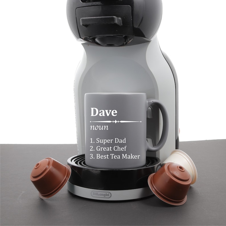 May include: A gray coffee mug with the name "Dave" and the definition "noun: 1. Super Dad 2. Great Chef 3. Best Tea Maker" printed on it. The mug is sitting on a gray coffee machine with two brown coffee pods.