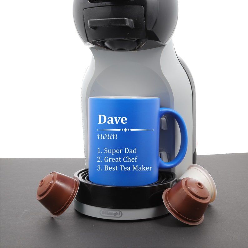 May include: Blue coffee mug with the word "Dave" and the definition "noun 1. Super Dad 2. Great Chef 3. Best Tea Maker" printed on it. The mug is sitting on a coffee machine.