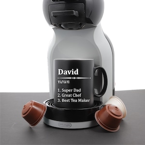 May include: Black coffee mug with white text that reads "David noun 1. Super Dad 2. Great Chef 3. Best Tea Maker". The mug is sitting on a gray coffee maker with a black base and a silver top. There are three brown coffee pods on the counter.