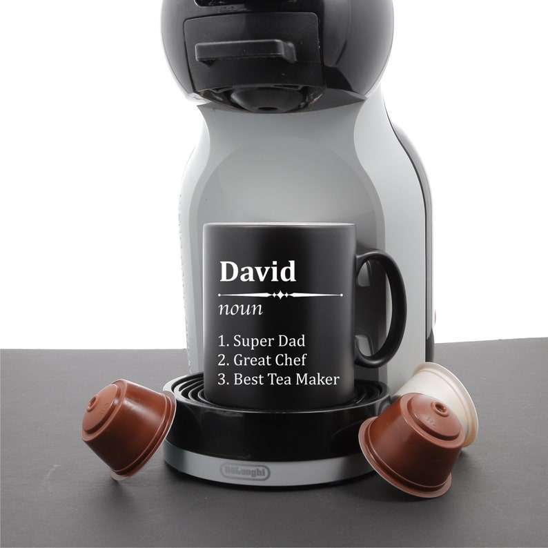 May include: Black coffee mug with white text that reads "David noun 1. Super Dad 2. Great Chef 3. Best Tea Maker". The mug is sitting on a gray coffee maker with a black base and a silver top. There are three brown coffee pods on the counter.