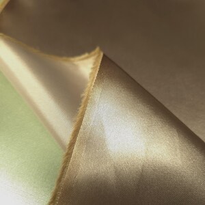 Puede incluir: Primer plano de una tela doblada en un tono marr&oacute;n dorado c&aacute;lido. La tela tiene una superficie lisa y brillante, con un ligero brillo. Los bordes de la tela son visibles, mostrando una textura cruda e inacabada. La tela parece ser un material lujoso, posiblemente para ropa o decoraci&oacute;n del hogar.