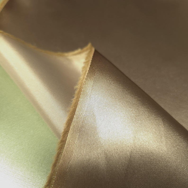 Puede incluir: Primer plano de una tela doblada en un tono marr&oacute;n dorado c&aacute;lido. La tela tiene una superficie lisa y brillante, con un ligero brillo. Los bordes de la tela son visibles, mostrando una textura cruda e inacabada. La tela parece ser un material lujoso, posiblemente para ropa o decoraci&oacute;n del hogar.