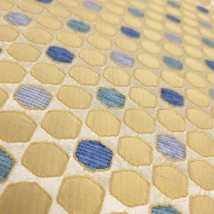 K&ouml;nnte beinhalten: Nahaufnahme eines Stoffes mit einem sich wiederholenden Muster aus gelben und blauen geometrischen Formen. Die Formen sind auf einen cremefarbenen Hintergrund gen&auml;ht. Die Textur des Stoffes ist sichtbar, mit leichtem Glanz.