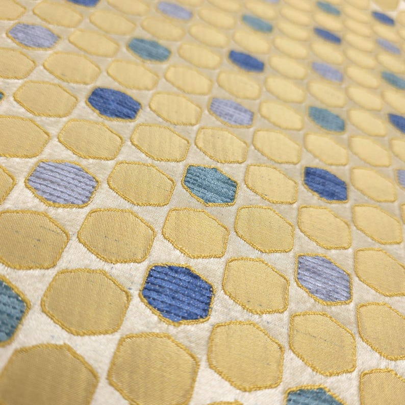K&ouml;nnte beinhalten: Nahaufnahme eines Stoffes mit einem sich wiederholenden Muster aus gelben und blauen geometrischen Formen. Die Formen sind auf einen cremefarbenen Hintergrund gen&auml;ht. Die Textur des Stoffes ist sichtbar, mit leichtem Glanz.