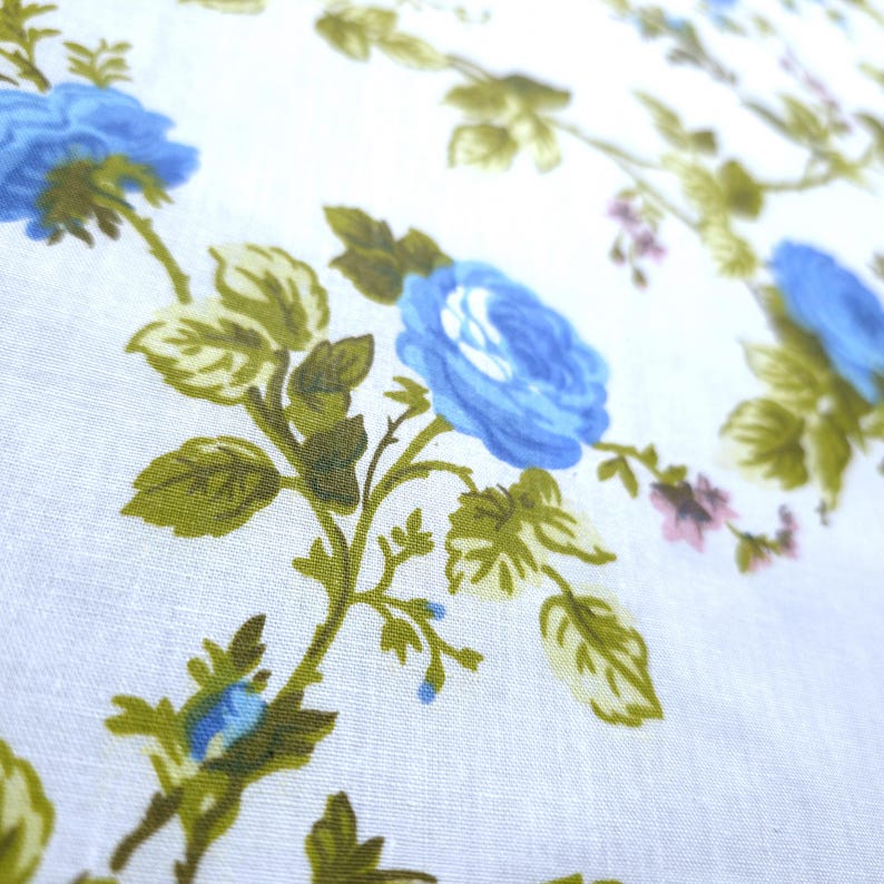 May include: Close-up of a white fabric with a floral pattern. The design features large, light blue roses with green leaves and stems. Small pink flowers are also scattered throughout the pattern.