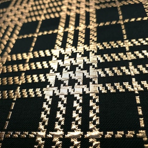 May include: Close-up of a black fabric with a gold houndstooth and plaid pattern. The fabric has a textured appearance with a repeating design of squares and geometric shapes. The gold color has a metallic sheen.