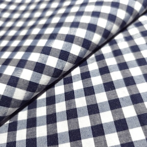 May include: Close-up of a folded fabric with a classic gingham pattern. The fabric features a repeating grid of navy blue, light blue, and white squares. The fabric is slightly rumpled, showing the texture and folds of the material.