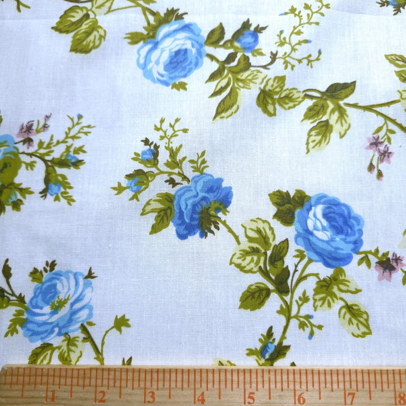 May include: White fabric with a repeating pattern of blue roses and green leaves. The roses are in full bloom and the leaves are detailed. A wooden ruler is at the bottom of the image, with inch markings.