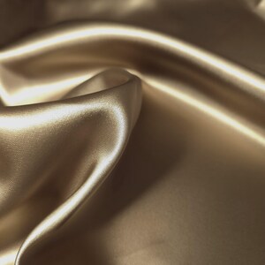 Puede incluir: Primer plano de una tela lujosa y fluida en un c&aacute;lido tono beige dorado. El material tiene una superficie lisa y brillante con pliegues y reflejos suaves, lo que sugiere una textura sedosa. La tela parece estar drapeada, creando una sensaci&oacute;n de movimiento y elegancia.