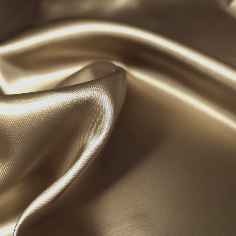 Puede incluir: Primer plano de una tela lujosa y fluida en un c&aacute;lido tono beige dorado. El material tiene una superficie lisa y brillante con pliegues y reflejos suaves, lo que sugiere una textura sedosa. La tela parece estar drapeada, creando una sensaci&oacute;n de movimiento y elegancia.