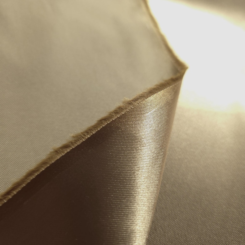 Puede incluir: Primer plano de una secci&oacute;n de tela en un tono marr&oacute;n dorado c&aacute;lido. El material tiene una textura suave, similar al sat&eacute;n, con un ligero brillo. El borde de la tela es visible, mostrando un borde en bruto sin terminar.