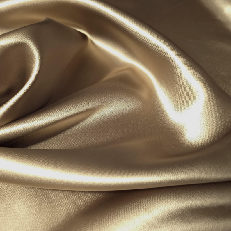 Puede incluir: Primer plano de una tela fluida de color beige dorado. El material presenta un brillo sedoso y brillante, con pliegues suaves y reflejos que crean una sensaci&oacute;n de profundidad y movimiento. La tela parece ser seda o sat&eacute;n.