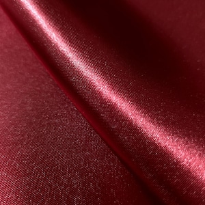 May include: Close-up of a deep red satin fabric. The fabric has a smooth, shiny surface with subtle texture and folds. The color is a rich, saturated shade of red, with highlights and shadows adding depth.