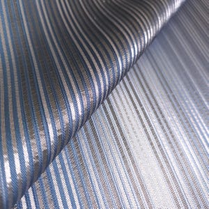 May include: A folded piece of fabric featuring a repeating pattern of vertical stripes in shades of light blue, dark blue, and metallic silver. The textile has a visible woven texture with a slight sheen, creating a striped design throughout.