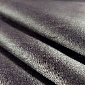 May include: Close-up of a dark gray fabric with a subtle sheen. The material is folded, creating deep shadows and highlights that emphasize the texture and weave of the textile. The fabric appears soft and luxurious.