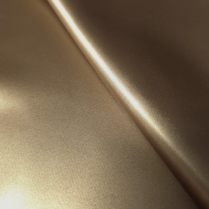 Puede incluir: Primer plano de una tela doblada de color marr&oacute;n dorado. El material tiene una textura sutil y un acabado brillante que refleja la luz. Los pliegues de la tela crean sombras y reflejos, sugiriendo un textil suave y lujoso.