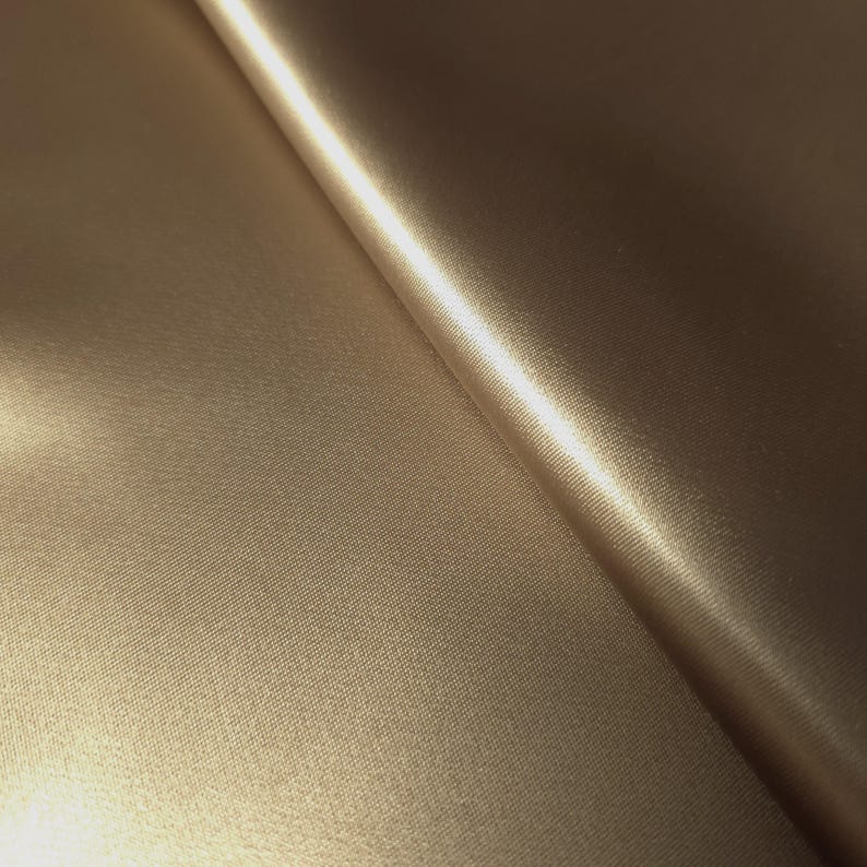 Puede incluir: Primer plano de una tela doblada de color marr&oacute;n dorado. El material tiene una textura sutil y un acabado brillante que refleja la luz. Los pliegues de la tela crean sombras y reflejos, sugiriendo un textil suave y lujoso.