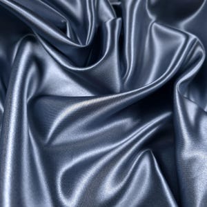 May include: Close-up of a flowing, lustrous fabric in a muted, silvery-blue hue. The material is tightly woven, with visible diagonal ribbing, and reflects light, creating highlights and shadows across its rippled surface. The fabric appears soft and pliable.