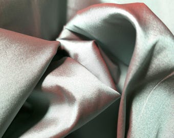 May include: Close-up of a folded fabric with a satin sheen. The fabric has a color gradient, transitioning from a light teal to a reddish-pink hue. The folds create shadows and highlights, emphasizing the fabric's texture.