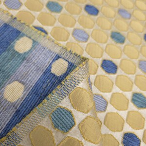 K&ouml;nnte beinhalten: Nahaufnahme eines gemusterten Stoffes mit einem sich wiederholenden Design aus gelben und blauen Formen auf wei&szlig;em Hintergrund. Der Stoff hat ein strukturiertes Aussehen, mit einer gefalteten Kante, die ein gestreiftes Muster in Blau und Wei&szlig; zeigt.