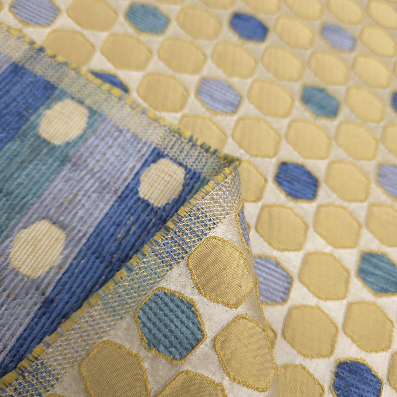 K&ouml;nnte beinhalten: Nahaufnahme eines gemusterten Stoffes mit einem sich wiederholenden Design aus gelben und blauen Formen auf wei&szlig;em Hintergrund. Der Stoff hat ein strukturiertes Aussehen, mit einer gefalteten Kante, die ein gestreiftes Muster in Blau und Wei&szlig; zeigt.