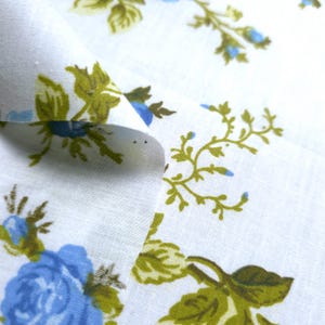 May include: Close-up of white fabric with a floral pattern. The design features blue roses and green leaves with a light blue accent. The fabric is folded, showing the texture and detail of the print.