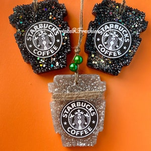 May include: Three Starbucks-themed air fresheners in the shape of coffee cups. Two are black with glitter, and one is clear with a twine accent. Each features the Starbucks logo. The background is orange.