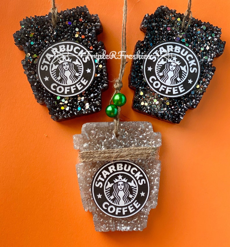 May include: Three Starbucks-themed air fresheners in the shape of coffee cups. Two are black with glitter, and one is clear with a twine accent. Each features the Starbucks logo. The background is orange.