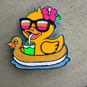 May include: A yellow duck wearing sunglasses and a pink flower, floating on a gold and blue pool float. The duck is drinking from a green cup with a straw. A small duck is on the left.