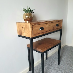 May include: A wooden console table with a single drawer and black metal legs. The table has a distressed wood finish and a gold plant pot on top. A smaller wooden shelf with black metal legs is attached to the bottom of the table.