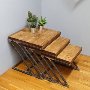May include: A set of three nesting tables with a wood top and black metal legs. The tables are arranged in a staggered pattern, with the largest table at the bottom and the smallest table at the top. The tables are decorated with two small potted plants.