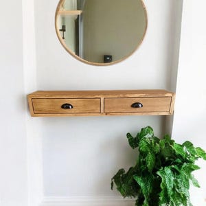 May include: A wooden floating shelf with two drawers and black metal handles. The shelf is mounted on a white wall, and a round mirror hangs above it. A green plant sits on the floor below the shelf.