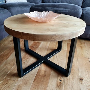 May include: A round wooden coffee table with a natural wood finish and black metal legs. The legs are in an X shape and the table is sitting on a wooden floor.