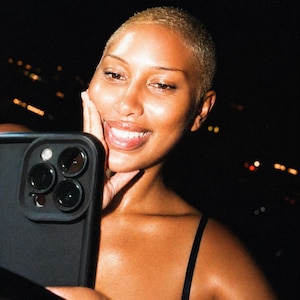 May include: A person with short blonde hair smiles while holding a black smartphone with three camera lenses. The person wears a black top, and the background is dark with blurred lights.