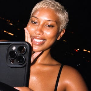 May include: A black smartphone with a triple-lens camera system held in a hand. The device features a matte black frame and a prominent square camera module containing three circular lenses and a flash, positioned against a dark background.
