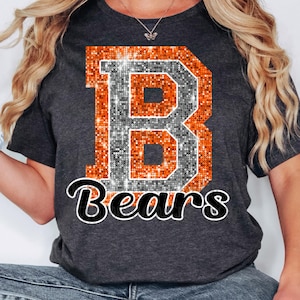 Può includere: T-shirt grigio scuro con una grande lettera "B" scintillante arancione e argento, e la parola "Bears" in corsivo bianco e nero. La maglietta è a girocollo.