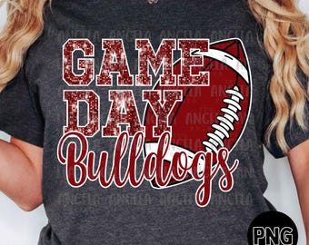 Peut inclure: T-shirt gris foncé avec un motif de football. Le t-shirt présente les mots "GAME DAY Bulldogs" dans une combinaison de paillettes rouges et de police script, ainsi qu'une illustration de ballon de football rouge et blanc.