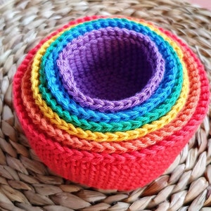 Peut inclure: Un ensemble de bols gigognes en crochet arc-en-ciel avec une couche intérieure violette. Les bols sont faits de fil et sont empilés les uns sur les autres.