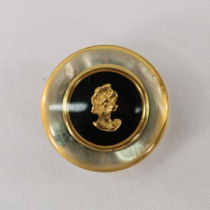 May include: A gold button with a black and white cameo of a woman's profile. The button has a white, pearlescent finish.