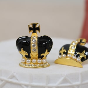Pode incluir: Duas coroas douradas com esmalte preto e detalhes de pérolas brancas. As coroas estão sobre uma superfície branca.
