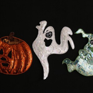 May include: Three sequined Halloween decorations on a black background. The decorations include an orange pumpkin with a carved face, a white ghost, and a green and blue ghost.
