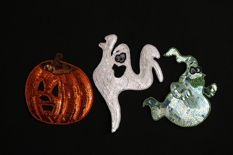 May include: Three sequined Halloween decorations on a black background. The decorations include an orange pumpkin with a carved face, a white ghost, and a green and blue ghost.
