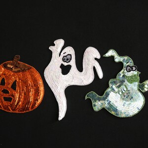 May include: Three Halloween-themed sequin patches on a black background. The patches include an orange pumpkin with a carved face, a white ghost, and a green ghost with a hat and glasses. These patches are likely for clothing or crafts.